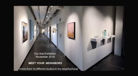 An exhibition hallway with art pieces displayed on walls and placard reading "our first exhibition november 2018 - meet your neighbors" with mention of artists from 16 local studios.
