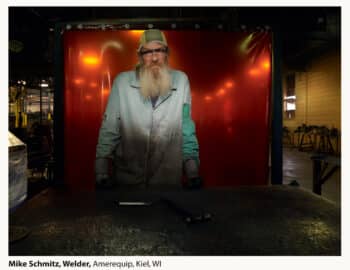 A welder named Mike Schmitz posing in his workshop in Kiel, WI, with work gloves and tools in front of a welding curtain lit with a red glow.