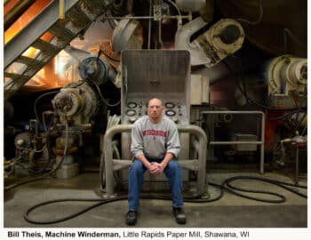 A man in a red shirt sits on a large metal chair in an industrial paper mill setting with machinery surrounding him.