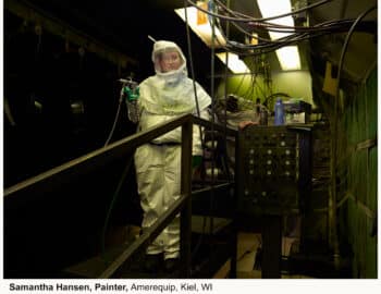 A painter in protective overalls holding a spray gun in an industrial setting with tangled cables and machinery around.