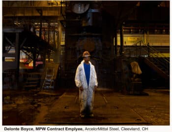 A worker in a white coverall and hard hat stands in the illuminated foreground of a dim, industrial steel mill.