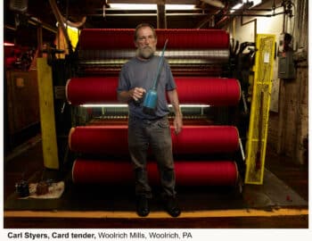 A man stands in a factory holding a mug, with red textile rolls behind him, at Woolrich Mills, Woolrich, PA. A caption identifies him as Carl Styers, a card tender.