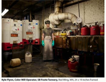 Man in a cap and apron stands in a tannery surrounded by large industrial dyeing machines and barrels, looking at the camera.