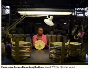 A woman sits at a workstation, displaying a decorative plate amidst stacks of similar plates, in a dimly-lit industrial setting.