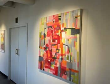 A colorful abstract painting with geometric shapes in pink, red, yellow, and gray hangs on a white wall next to a closed set of double doors.