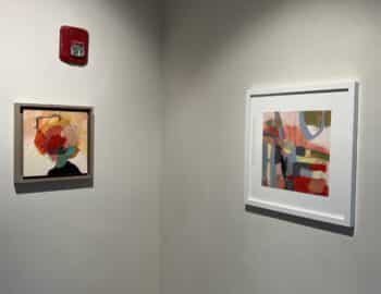 Two abstract paintings on a wall, one with a wooden frame depicting colorful shapes, and the other in a white frame featuring geometric patterns. A red fire alarm is above the left painting.