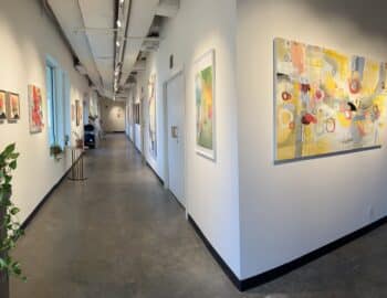 Art gallery hallway with abstract paintings on white walls and a concrete floor. Potted plant visible on the left.